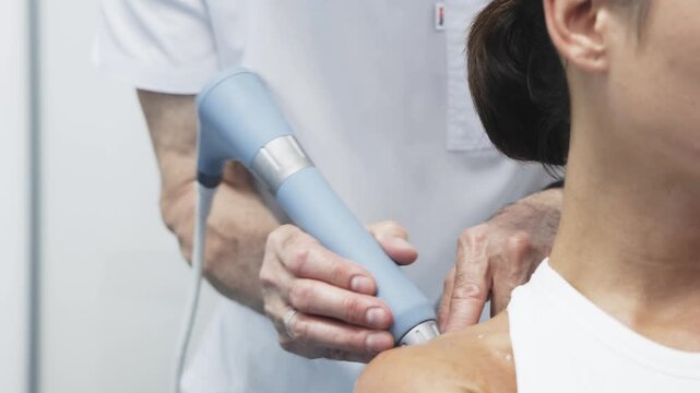 Therapist applying shock wave therapy device to the shoulder of a female patient, showcasing the treatment process in a clinical setting with visible equipment and professional attire