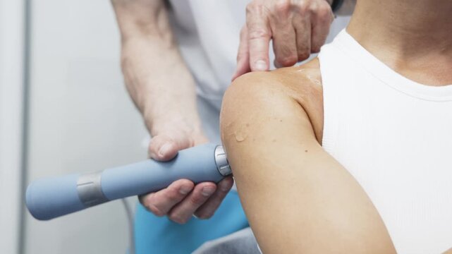 Female patient receiving shock wave therapy on shoulder by male therapist in clinical setting, focusing on treatment device and shoulder area during procedure