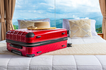 Naklejka premium Red suitcase and straw hat on a bed in a luxury hotel room with panoramic glass windows overlooking mountains and a blue sky.