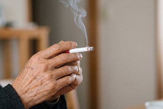 Wrinkled hand of an elderly man holding a lit cigarette with smoke