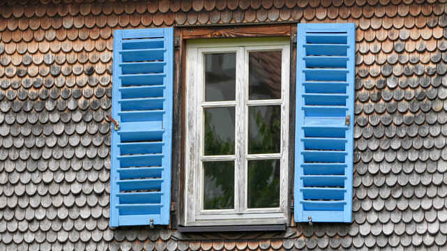 traditional window with bright blue wooden shutters on a wall with wooden shingles