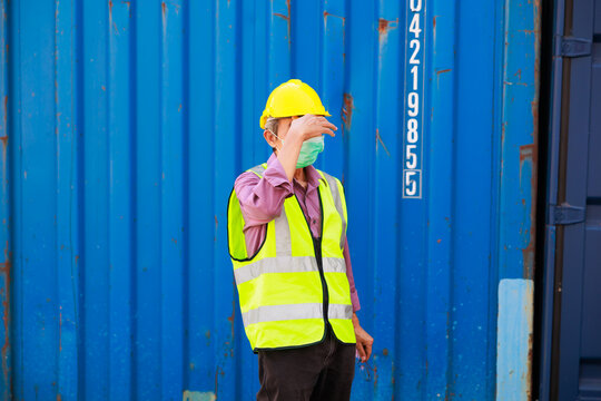 Headahe, Tired asian senior male container worker in protective face mask, hardhat helmet and uniform resting at container yard port of import and export.