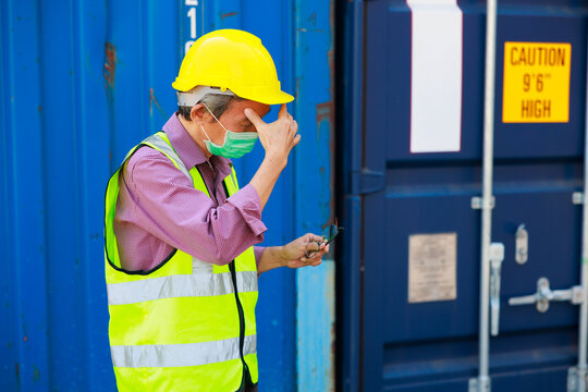Headahe, Tired asian senior male container worker in protective face mask, hardhat helmet and uniform resting at container yard port of import and export.