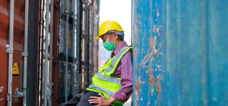 Tired asian senior male container worker in protective face mask, hardhat helmet and uniform resting at container yard port of import and export.