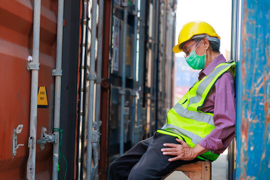 Tired asian senior male container worker in protective face mask, hardhat helmet and uniform resting at container yard port of import and export.