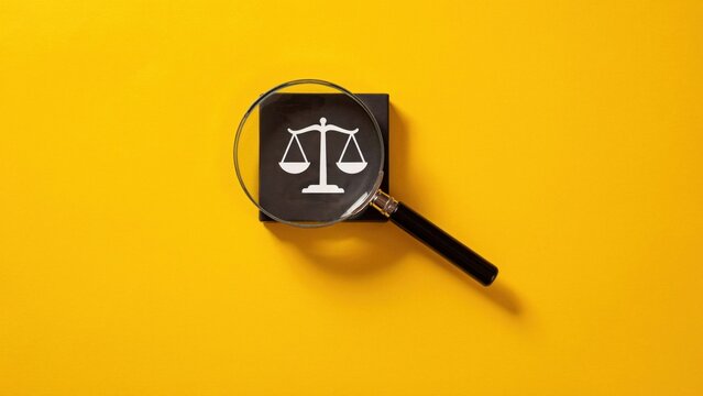 Magnifying glass examines scales of justice on a yellow background closely.
