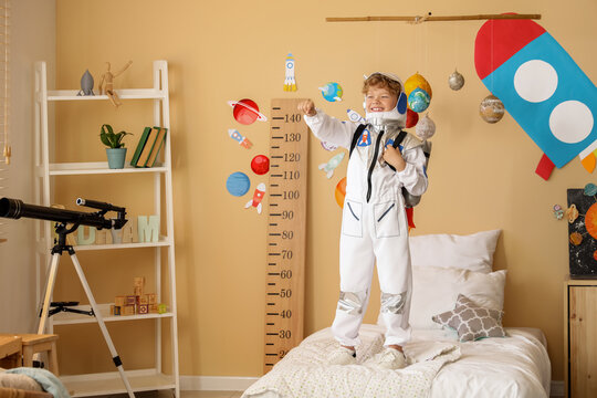 Happy young boy in astronaut costume standing on bed with space-themed room decor and telescope