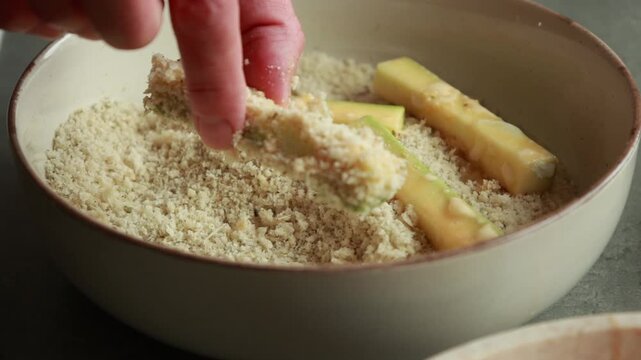 Close-up of egg-washed zucchini fries being rolled and generously coated in a crunchy panko breadcrumb and parmesan cheese mixture