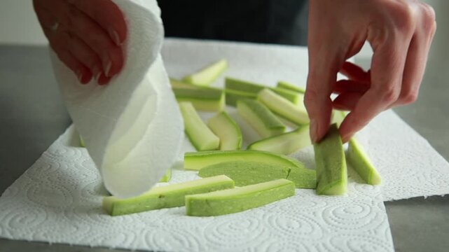 Close-up of a woman's hands gently patting raw zucchini fries with a white paper kitchen towel to remove excess moisture before breading