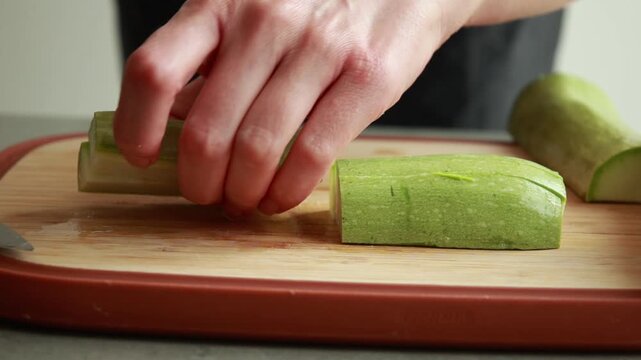 Close-up of a woman's hands slicing a fresh raw zucchini into uniform fry-shaped sticks on a wooden cutting board