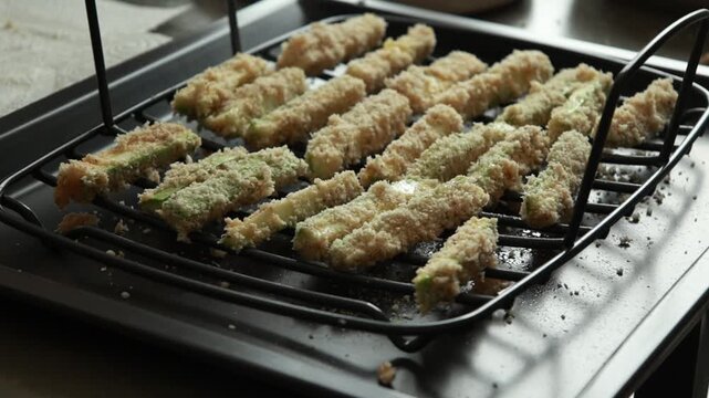 Close-up of raw zucchini fries coated in parmesan panko breadcrumbs and spices, neatly arranged on a wire rack ready for baking