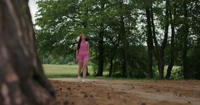 Attractive sporty girl in pink romper strolling among trees in summer park