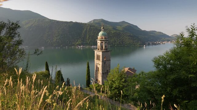 Ancient church bell tower in Morcote village overlooking the calm lake water. Stabilizer shot of historical stone architecture in Ticino canton of Switzerland. Morcote landmark in Switzerland