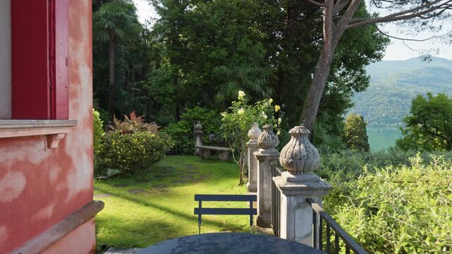 Cozy villa terrace inside Parco Scherrer in Morcote. Beautiful green garden with roses and palms surrounds stone balustrade next to Lake Lugano. Parco Scherrer botanical park, Morcote, Switzerland