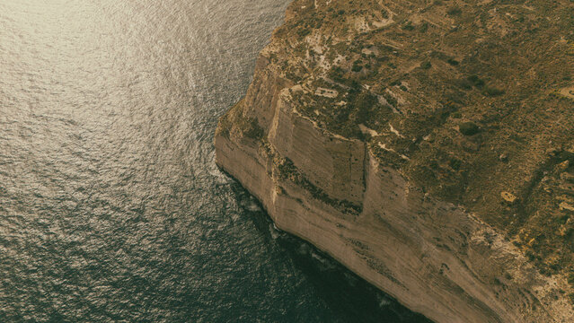 high angle drone view looking down at dingli cliffs chalky limestone coast on south malta island coast with Mediterranean Sea below at summer sunset golden haze above with green foliage on the land