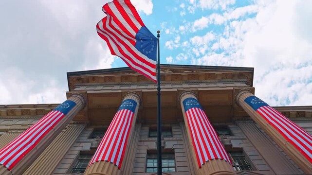 Low angle view of patriotic banners on government building, Washington D.C. Columns wrapped in red, white, and blue fabric with flag waving against cloudy sky.