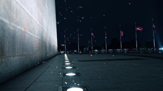 Ground level view of Washington Monument base at night, Washington, D.C. Row of uplights along marble wall with American flags on poles and insects in beam glow.