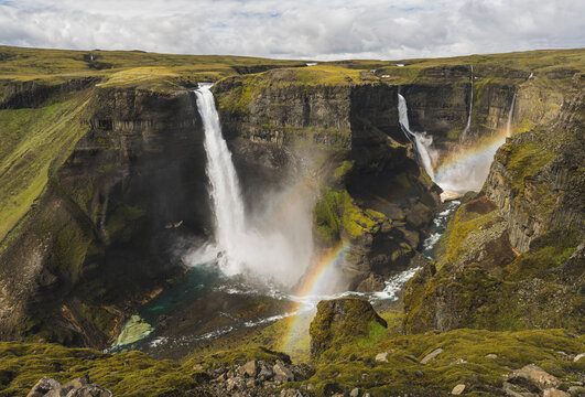 Spectacular double waterfall dropping into a steep green canyon with vivid rainbows forming in the mist.