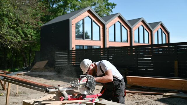 Builder trims timber plank with circular miter saw in bright daylight. Carpentry work produces fine sawdust during material preparation and exterior installation