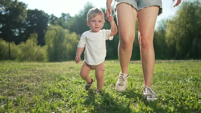 Mom helps toddler walk on green grass in park. Toddler exploring nature during first steps with mom. Joyful moment as toddler and mom walk on grass. Toddler learning to walk on grass with mom support.