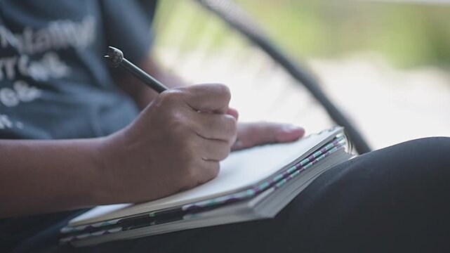 Close up of young man hands holding pen and writing text on blank paper notebook outdoor. Female taking notes, planning, studying or writing a diary in nature background during day
