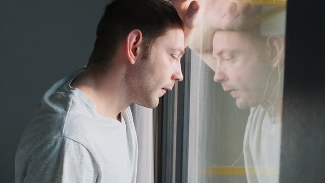 Thoughtful man looking through window with reflection feeling loneliness and depression at home. Male in quiet moment of sadness and self reflection, suggesting midlife crisis, emotional stress