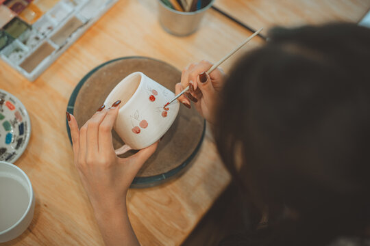 Close up of Asian woman painting cherry pattern on ceramic mug with paintbrush, creative female artisan handcrafting pottery cup at table in studio workshop for hobby, small business, and handmade con