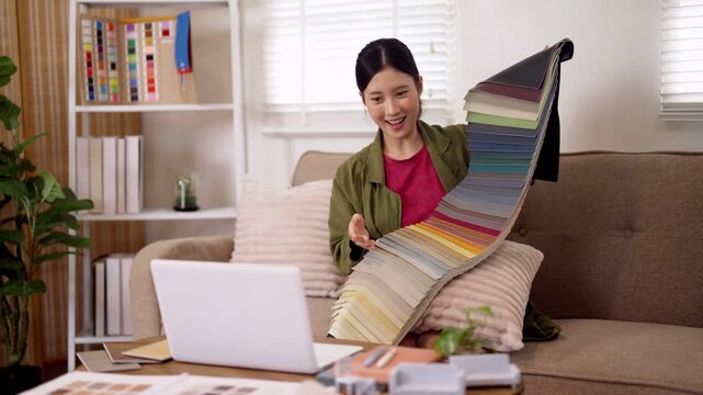 Young Asian interior designer presenting colorful fabric swatch palette to camera while consulting with laptop in modern bright home office space