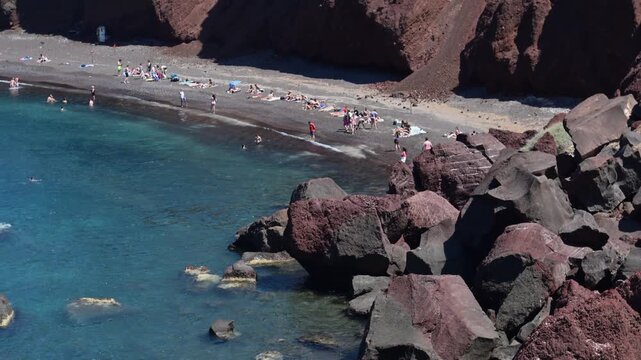 Santorini, Greece - 11.06.2026: Visitors relax and swim along a dark volcanic shoreline with red cliffs and clear Aegean water.