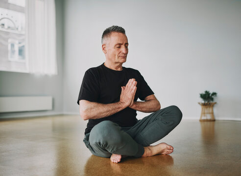 Mature man sitting with his eyes closed and meditating in the lotus pose while sitting alone on the floor of a yoga studio