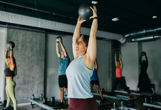 Mature woman in sportswear holding a ball and stretching her arms in the air while standing on a reformer during an exercise class at a pilates gym