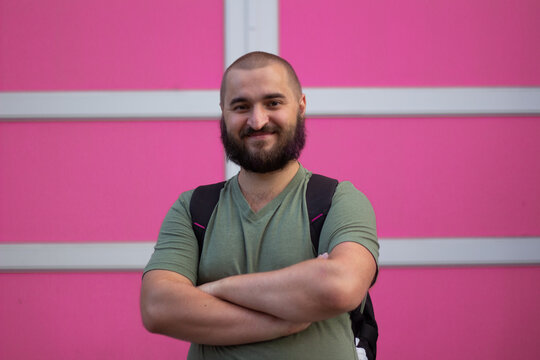 Street city photo. Young adult bald white man with beard, green t-shirt, black backpack, crossed his arms, stands near pink house wall, looks at camera. Concept of tourism, travel, trip, life, rest