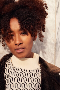 Portrait of a confident young African American woman with curly hair wearing a geometric sweater and brown coat outdoors.