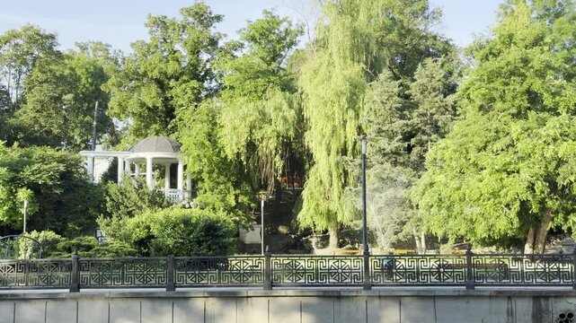 A beautiful white rotunda stands surrounded by lush green trees. The sunny park embankment feels incredibly peaceful, calm, and charming.