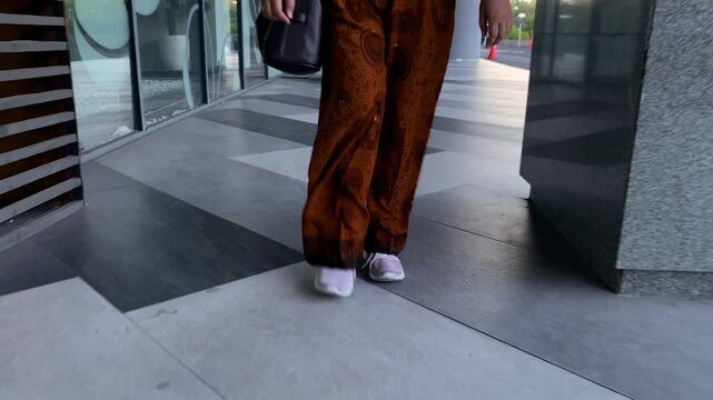 Happy fashionable stylish adult Asian woman wearing batik traditional long pants and outer knitted jacket walking through modern office building hallway and looks at wristwatch.