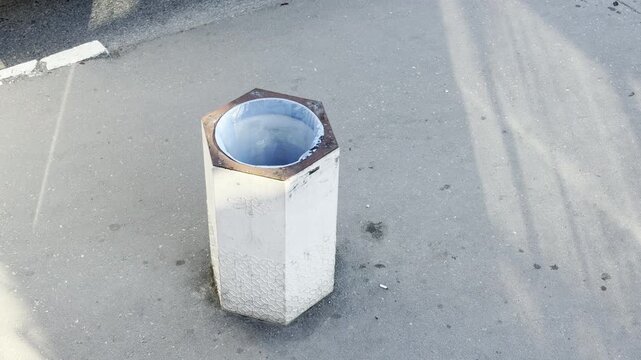 A white hexagonal trash bin stands orderly on a sunny asphalt pavement. The clean and quiet setting brings a peaceful, serene feeling to the empty urban space.