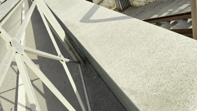 A smooth stone ledge and a metal fence sit under the bright sun near a city stairway. The empty space feels calm yet stark. Crisp shadows create a lonely, quiet mood.