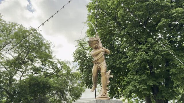 A golden cherub statue spouts a stream of water in a lush green park. Hanging strings of lights add a whimsical touch. The charming scene evokes a peaceful and joyful mood.