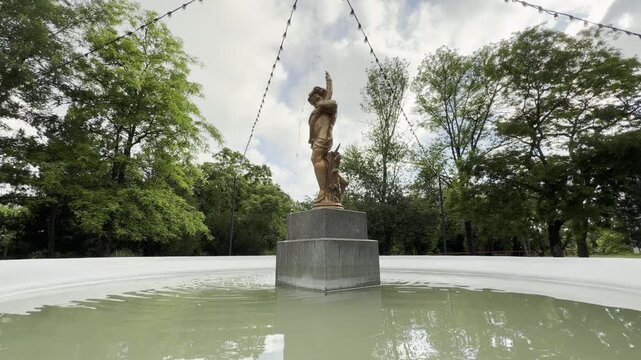 A golden cherub statue spouts water inside a circular park fountain pool. Lush green trees and string lights surround the water, creating a whimsical and deeply peaceful mood.