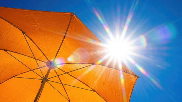 Bright sun flare over orange beach umbrella in clear blue sky, excessive heat, heatwave global warming