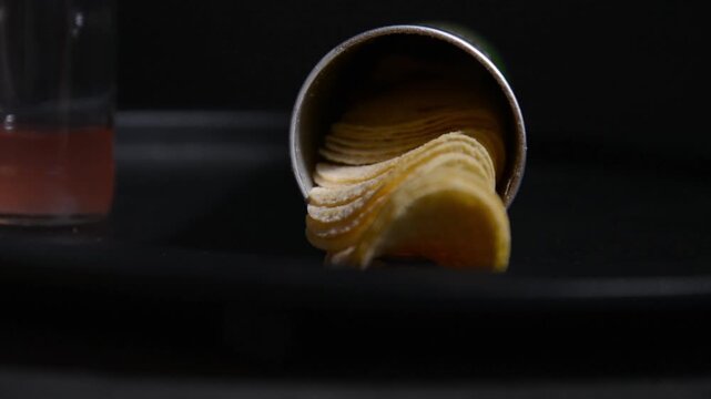 Close up of a cylindrical chip container lying on a surface while a hand repeatedly takes potato chips from it. Snack scene focused on food, texture and consumption.
