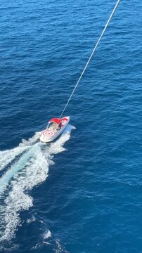 Speedboat creates a white wake while towing a parasail rider above the open sea. Water sports tourism, coastal recreation, summer holiday, marine adventure, aerial leisure experience.