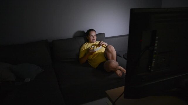 Wide shot of a young woman lying on a sofa and eating potato chips while watching a movie. The back of a television is partially visible in the frame, creating a relaxed home entertainment scene.