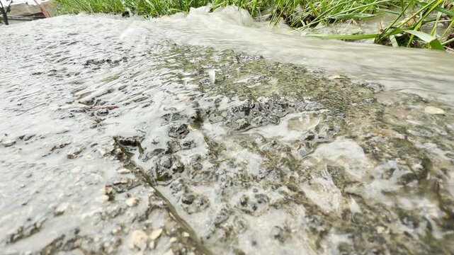 heavy floodwater flows rapidly over rough asphalt. The surging current splashes forcefully over the road edge into the grass, creating a loud rushing sound.