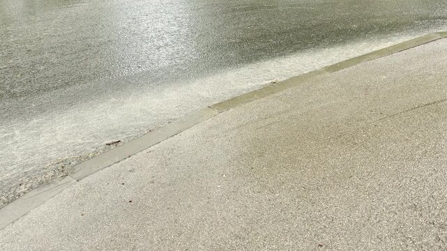 a stream of murky rainwater surges quickly down the edge of an asphalt road. The fast current flows against the concrete curb, creating a steady rushing and splashing sound.