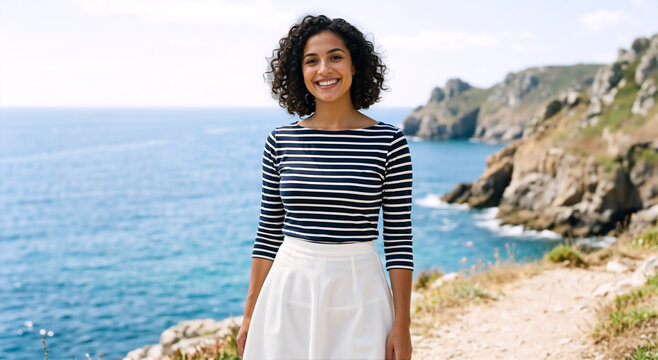 Smiling young woman with curly hair standing on a coastal path. Happy female tourist in a striped shirt overlooking the ocean. Travel and seaside vacation concept