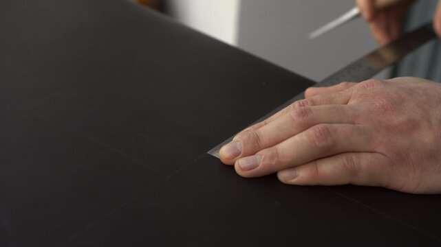 Leather craftsman tracing a pattern using an awl and a metal ruler on a black leather piece to create a custom product