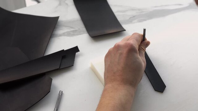 Artisan's hands punching holes in a black leather strip using a hammer and a hole punch tool on a white cutting board in a workshop