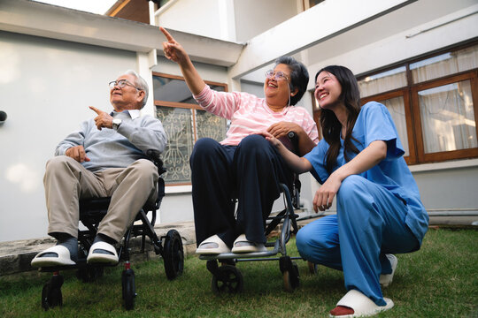 Asian nurse with senior couple wheelchairs pointing finger happy elderly retired husband wife pointing up garden, smiling nurse kneeling beside senior partners sitting wheelchairs backyard.
