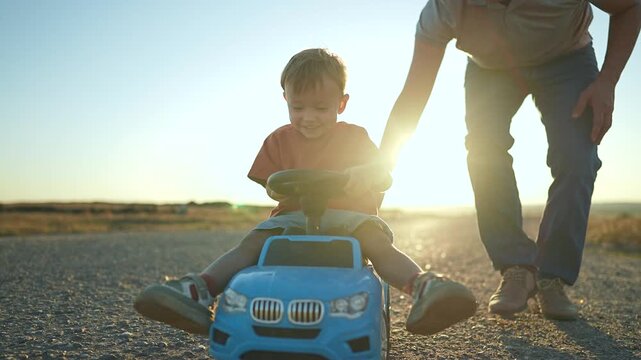 Father pushes kid on toy car outdoors. Father and kid play together during sunset. Fun family time with toy car. Kid enjoys ride while father helps. Father bonds with kid through playful car ride.